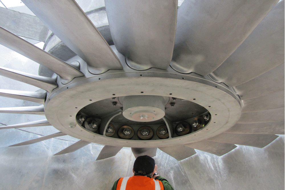 A man in an orange hi vis vest making adjustments to the huge axial fan he is installing over head
